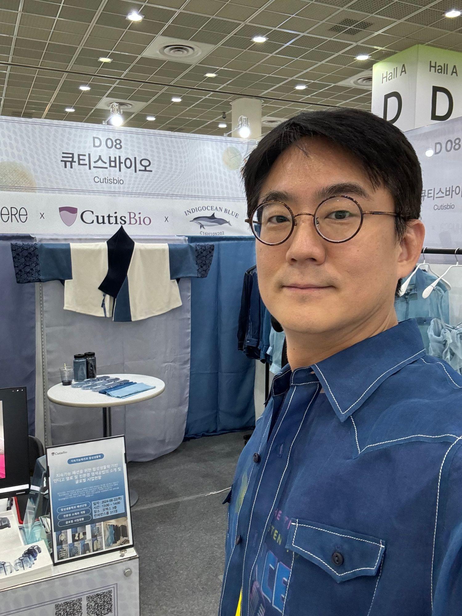 展示会の CutisBio ブース(D08)の前で、バイオインディゴで染めたシャツを着ている様子。後ろに染色サンプル布とインディゴ粉末の瓶が見える